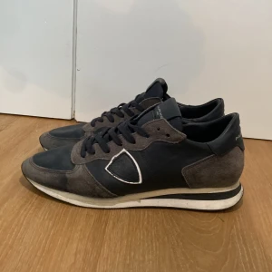 Philippe Model gråblå sneakers - Snygga sneakers från Philippe Model i en mörkblå och grå färgkombination. Skorna har klassisk snörning, rund tå och detaljer i mocka och skinn. Vit och svart randig sula ger en sportig vibe. Perfekt för dig som gillar stilrena och exklusiva sneakers.