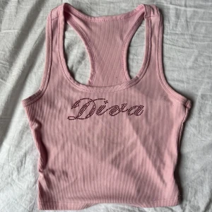 Rosa ribbad linnetopp från SHEIN - Säljer en ljusrosa ribbad linnetopp från SHEIN i storlek M. Toppen har racerback och ärmlös design, med glittriga röda stenar som bildar texten 'Diva' framtill. Materialet är mjukt och stretchigt polyester. Perfekt för dig som vill sticka ut med en cool och feminin vibe.
