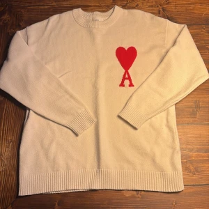 Vit stickad tröja från Ami Paris - Säljer en Vit stickad tröja från Ami Paris ANVÄND 5 GÅNGER, med röd hjärtlogga och bokstaven A på bröstet. Tröjan har rund halsringning, ribbade muddar och är långärmad. Perfekt för dig som gillar stilrena plagg med en twist.