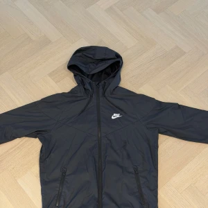 Svart Nike vindjacka med huva - Snygg svart vindjacka från Nike med huva och hel dragkedja framtill. Jackan har två fickor med dragkedja och meshfoder på insidan. Perfekt för blåsiga dagar och enkel att matcha med streetwear. Klassisk Nike-logga på bröstet.