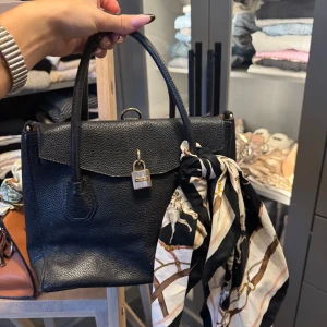 Svart Michael Kors väska med scarf och kuvertväska - Stilren svart handväska i skinn med guldfärgade detaljer och lås framtill. Medföljer matchande svart kuvertväska med samma låsdetalj. Handväskan har en snygg scarf knuten runt handtaget och flera fack inuti. Perfekt accessoar för en trendig look.