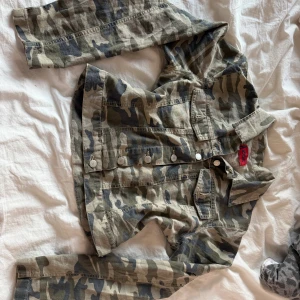 Camouflage jeansjacka från ZOE - Snygg camouflagefärgad jeansjacka från ZOE med klassisk krage, knappar framtill och två bröstfickor. Jackan har lång ärm och är tillverkad i slitstarkt bomullsmaterial. den är i stl xs/s. knappt använd men köpt på second hand