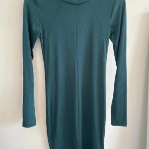 Mörkgrön ribbad klänning Gina Tricot - Snygg mörkgrön ribbad klänning från Gina Tricot med lång ärm och rund hals. Modellen är kroppsnära och tillverkad i stretchigt material med 95% polyester och 5% elastan. Perfekt för dig som gillar enkel och stilren design.