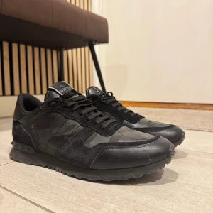 Valentino Rockrunner svarta sneakers - Svarta Valentino Rockrunner sneakers med coola nitar på hälen och diskret camouflagemönster. Skorna har en mix av mocka och skinn, snörning och en chunky sula. Perfekta för dig som gillar streetstyle med lyxig touch. 