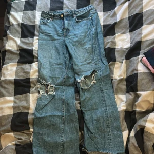 Blå jeans med slitna knän från 157 - Säljer ett par blå jeans från 157 i storlek M. Byxorna har breda ben och coola slitningar vid båda knäna för en edgy look. Klassisk femficksmodell med knapp och dragkedja framtill. Perfekt för dig som gillar streetstyle.