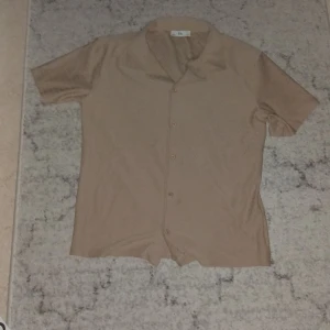 Beige kortärmad skjorta från C&A - Snygg beige skjorta från C&A med korta ärmar och klassisk krage. Skjortan har knappar framtill och ett diskret fiskbensmönster i tyget som ger en cool touch. Perfekt för dig som gillar stilrena och enkla plagg.