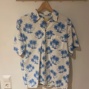 Rotation kortärmad skjorta med palmer - Säljer en vit kortärmad skjorta från Rotation med blått palmtryck. Skjortan har klassisk krage, knappar framtill och bröstficka. Perfekt för sommarens chill dagar. Materialet känns lätt och luftigt.