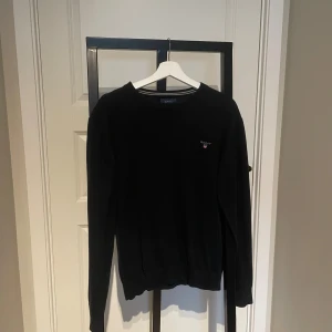 Svart stickad tröja från Gant - Klassisk svart stickad tröja från Gant med rund halsringning och diskret broderad logga på bröstet. Tröjan har ribbade muddar och är långärmad, perfekt för lager-på-lager-stil under kyligare dagar.