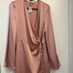 Rosa omlottklänning i satin från H&M - Superfin kort rosa omlottklänning i satin från H&M. Klänningen har långa ärmar, djup v-ringning och knyt i midjan för en snygg siluett. Perfekt för dig som gillar en stilren och trendig look.
