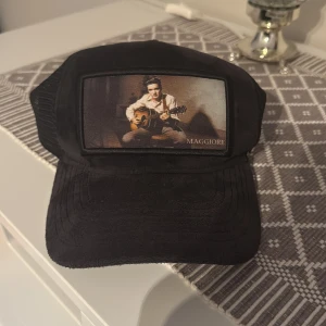 Svart keps från Maggiore med tryck - Svart keps från Maggiore med en stor patch framtill som visar en person med gitarr. Kepsen har mesh på insidan och justerbar baksida. Snygg och unik detalj med Maggiore-logga på insidan. Perfekt för dig som vill sticka ut.