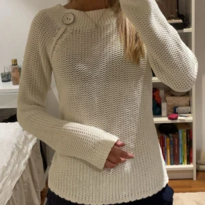 Vit stickad tröja med knappdetalj - Säljer en vit stickad tröja med lång ärm och en stor dekorativ knapp vid axeln. Tröjan har en lite bredare halsringning och är mjuk och skön, perfekt för kyligare dagar.