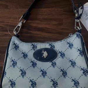 Blå axelväska från U.S. Polo Assn. - Snygg mini axelväska från U.S. Polo Assn. i blått  med polospelare-mönster. Väskan har silverfärgad logga framtill, dragkedja och avtagbar axelrem. Ny och oanvänd!! 🩵🩵 Öppen för prisförslag!