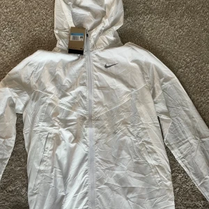 Vit Nike vindjacka med huva M - Nike Running vindjacka i vitt med diskret prickmönster, hel dragkedja och huva. Jackan är lätt, har fickor med dragkedja och kan packas ihop smidigt. Swoosh-logga på bröstet och detaljer på insidan. Perfekt för löpning och aktiva dagar.