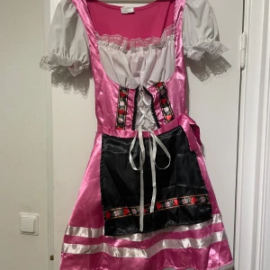 Rosa och vit dirndlklänning Boland M - Superfin rosa och vit dirndlklänning från Boland med puffärmar, spetsdetaljer och svart förkläde. Klänningen har snörning framtill, blommigt band och glansigt tyg. Perfekt för temafest eller oktoberfest! Sidenliknande känsla och klassisk tysk stil.