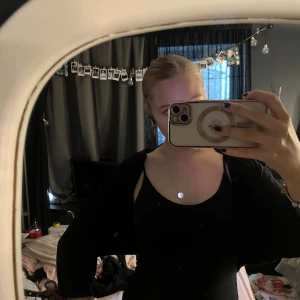 Svart croppad kofta med lång ärm - En svart, tunn och croppad kofta med lång ärm. Koftan är öppen framtill och har en enkel, stilren design som passar perfekt över linnen eller klänningar. Materialet känns mjukt och lätt, vilket gör den skön att bära under både vår och sommar.