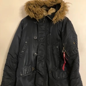 Blå parkajacka Alpha Industries M - Parkajacka från Alpha Industries i blått med fluffig brun fuskpäls på huvan och orange foder. Jackan har flera fickor, stora knappar, dragkedja och den klassiska 'Remove Before Flight'-taggen på ärmen. Perfekt för kalla dagar med sin rejäla och varma design.