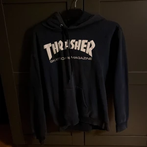 Thrasher hoodie - Säljer en mörkblå Thrasher hoodie med klassiskt vitt logotryck på bröstet. Tröjan har huva med snörning och en stor magficka. Perfekt för dig som gillar streetwear och skatekänsla. Storlek S