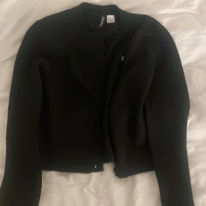 Svart kofta från H&M Divided XS - En stilren svart kofta från H&M Divided i storlek XS. Koftan har rund halsringning, lång ärm och en minimalistisk look utan synliga knappar. Perfekt att slänga över en topp för en clean vibe.