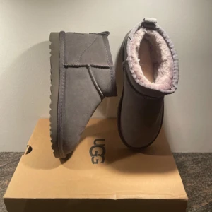 Grå UGG boots med fårskinn - Säljer ett par grå UGG boots med rund tå och platt sula. Skorna är fodrade med mjukt fårskinn på insidan och har en klassisk UGG-logga baktill. Yttermaterialet är mocka och de har en låg, robust siluett som är perfekt för kalla dagar. Storlek 37 helt nya aldrig använda   Box ingår ej i köp av dem