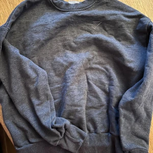 Mörkgrå sweatshirt från Gymshark - Mörkgrå sweatshirt från Gymshark med diskret logga på bröstet. Klassisk rund halsringning och ribbade muddar vid ärmslut och nederkant. Perfekt för en chill och sportig look.