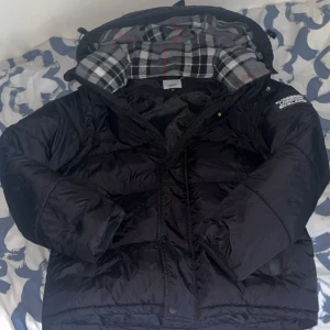 Svart pufferjacka från Burberry - Säljer en svart pufferjacka från Burberry med klassiskt rutigt foder i huvan. Jackan har dragkedja, tryckknappar och en tydlig Burberry London England-logga på ärmen. Perfekt för kalla dagar och riktigt snygg streetstil.