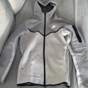 Grå Nike Tech med huva XS - Säljer en grå Nike tech från Nike i storlek XS. Jackan har hel dragkedja, huva och en svart detalj längs bröstet. Ficka med dragkedja på ena ärmen och diskret Nike-logga på bröstet. Perfekt för dig som gillar sportig och clean stil.