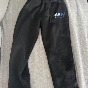 Svarta mjukisbyxor från Trapstar - Svarta mjukisbyxor från Trapstar med broderad logga i blått och vitt på vänster ben. Byxorna har snörning i midjan och dragkedjor vid bensluten. Perfekta för en chill och avslappnad stil.