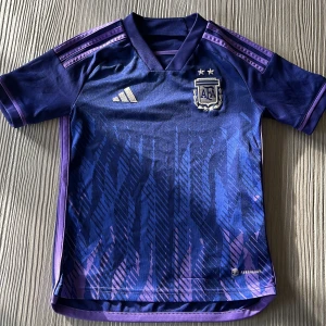 Argentina bortatröja Adidas 9-10 år - Säljer en officiell Argentina bortatröja från Adidas i mörkblått och lila med guldiga detaljer. Tröjan har korta ärmar, broderat AFA-märke och klassiska Adidas-ränder på axlarna. Tillverkad i lätt och ventilerande polyester med Aeroready-teknologi. Den är även äkta.