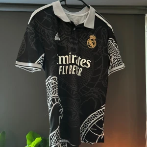 Real Madrid svart draktröja Adidas S - Säljer en svart Real Madrid fotbollströja från Adidas i storlek S. Tröjan har coolt drakmönster i grått, vit krage och detaljer samt klubbmärke i guld. Tryck med Emirates Fly Better på bröstet och nummer 13 med namn på ryggen. Perfekt för dig som älskar unika matchtröjor.                          Tyvärr har jag mitt efternamn på ryggen…