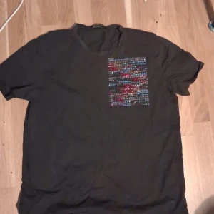 Svart Just Cavalli t-shirt med paljettdetalj - Svart t-shirt från Just Cavalli i storlek XL. Framsidan har en fyrkantig detalj med färgglada paljetter i blått, rött och silver. Klassisk rund halsringning och korta ärmar. Perfekt för dig som vill sticka ut med en unik detalj.