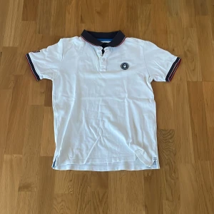 Vit Sail Racing pikétröja med emblem - Vit piké från Sail Racing med marinblå krage och röd/vit kant. Kortärmad modell med amerikansk flagga på ärmen och Sail Racing Performance Technology-emblem på bröstet. Snygga detaljer och klassisk look, perfekt för dig som gillar sportig stil.