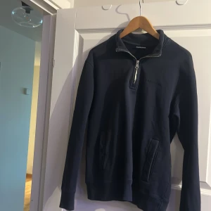 Mörkblå half zip tröja J.Lindeberg - Mörkblå långärmad tröja från J.Lindeberg i storlek S. Tröjan har en snygg half zip-dragkedja vid halsen och broderad logga på bröstet. Tillverkad i mjuk bomull med två fickor framtill. Perfekt för en stilren och avslappnad look.