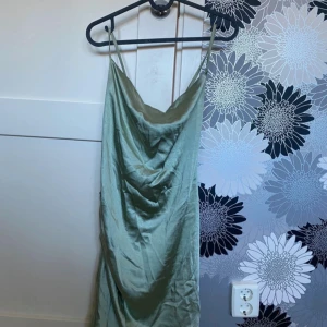 Ljusgrön klänning från NA-KD - Snygg ljusgrön klänning från NA-KD med tunna axelband och draperad halsringning. Klänningen har en mjuk satinliknande yta och knytband i ryggen för extra detalj. Perfekt för dig som vill ha en chic och trendig look.