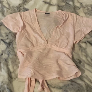 Ljusrosa omlottopp med knyt - Säljer en ljusrosa omlottopp från Gina Tricot med korta ärmar och v-ringning. Toppen har knytband i midjan som ger en snygg siluett och är tillverkad i mjuk bomull. Perfekt för dig som gillar en feminin och chill stil. aldrig använd