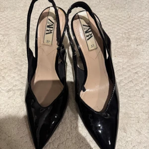 Svarta klackar  Zara,  37 - Snygga svarta pumps från Zara i storlek 37. Skorna har spetsig tå, smal klack och är gjorda i glansigt lackmaterial. Slingback-modell med öppen häl och elegant form som ger en stilren look.