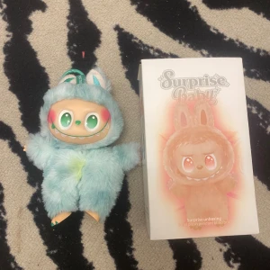 Surprise Baby Vinyl Plush Pendant - Söt Surprise Baby vinylfigur i mjuk blå dräkt med gröna ögon och rosa kinder. Levereras med originalförpackning. Figuren är i gott skick, med mindre missfärgning på magen. Perfekt som samlarobjekt eller present.