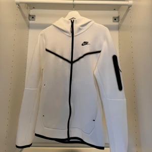 Vit Nike hoodiejacka med svarta detaljer - Nike hoodiejacka i vitt med svarta dragkedjor och linjer. Jackan har huva, hel dragkedja framtill och en extra ficka med dragkedja på ärmen. Snygg sportig look och skön passform, perfekt för chill eller träning.