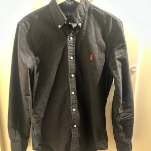 Svart skjorta från Ralph Lauren - Svart skjorta från Ralph Lauren med klassisk button-down krage och vita knappar. Ikonisk röd broderad logga på bröstet. Skjortan är långärmad och har en normal passform, tillverkad i mjuk bomull som känns skön mot huden.                             Bara att skriva vid frågor och funderingar.  Priset är ej hugget i sten 