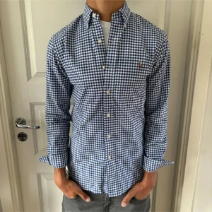 Blårutig skjorta från Ralph Lauren - Snygg blårutig skjorta från Ralph Lauren i slim fit-modell. Skjortan har klassisk krage, knappar framtill och det ikoniska broderade Polo-logot på bröstet. Tillverkad i stretchig bomull som sitter skönt och ger en fräsch look. Perfekt för dig som gillar stilrena detaljer.