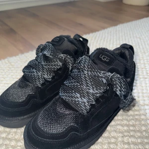 Svarta UGG sneakers med pälsfoder - Snygga svarta sneakers från UGG med grova, mönstrade snören och fluffigt pälsfoder på insidan. Ovandelen är i mocka och textil, med en robust platt sula och rund tå. Perfekt för dig som vill ha både stil och komfort under höst och vinter.