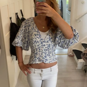 Blommig blus med puffärm från H&M - Superfin blus från H&M i linneblandning med blått blommönster på vit botten. Modellen har v-ringning, knappar framtill och stora puffärmar som ger en trendig look. Perfekt till ljusa jeans.