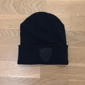 Svart DIF mössa med broderad logga - Svart ribbstickad mössa från Djurgårdens IF med uppvikt kant och broderad DIF-sköld framtill. Klassisk och stilren modell, perfekt för kalla dagar. Officiell supporterprodukt.