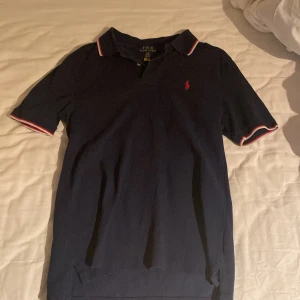 Marinblå pikétröja från Ralph Lauren - Säljer en klassisk marinblå pikétröja från Ralph Lauren med röd broderad logga på bröstet. Tröjan har korta ärmar och krage med vita och röda detaljer på ärmslut och krage. Perfekt för en stilren och sportig look.