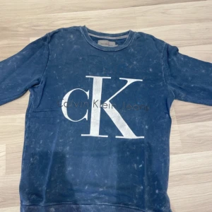Blå sweatshirt från Calvin Klein Jeans - Blå sweatshirt från Calvin Klein Jeans med stort vitt CK-tryck på bröstet. Tröjan har rund hals, långa ärmar och ribbade muddar. Snygg detalj med svart läderpatch på ärmslutet. Tillverkad i mjuk bomull, perfekt för en avslappnad stil.