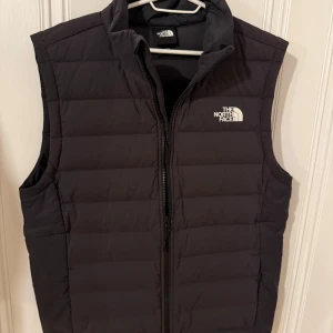 Svart dunväst från The North Face - Säljer en svart dunväst från The North Face i storlek M. Västen har en quiltad design, hög krage och dragkedja framtill. Perfekt för lager-på-lager och håller dig varm under kyliga dagar. Klassisk logga på både bröstet och ryggen.
