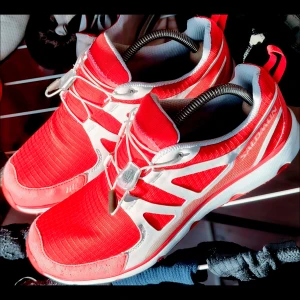 Salomon Sneakers/Löparskor Unisex [40+2/3]  - Säljer ett par sportiga röda och vita Salomon sneakers med snabbsnörning och slitstark platt sula. Skorna har en modern design med vita detaljer och mesh-material som andas. Perfekta för dig som gillar att sticka ut med färg och vill ha bekväma skor till träning eller vardag. Finns spår av bruksslitage på en av skornas hätta men knappt sebart. 