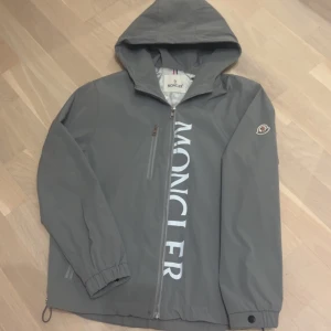 Grå vindjacka med huva från moncler - Snygg grå vindjacka från Moncler med huva och stor vit logga längs dragkedjan. Jackan har elastiska muddar, bröstficka med dragkedja och klassisk Moncler-patch på ärmen. Perfekt för dig som gillar stilren och sportig look.