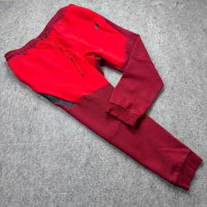 Röda Nike mjukisbyxor 3XL - Nike mjukisbyxor i rött och vinrött med svart logga på vänster lår. Byxorna har dragsko i midjan, muddar vid benslut och en svart detalj vid fickan. Perfekta för chill eller träning, tillverkade i mjukt bomullsmaterial.