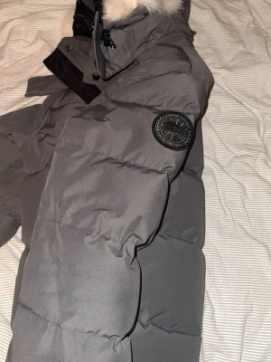 Grå pufferjacka från Canada Goose - Säljer en grå pufferjacka från Canada Goose med svart patch på ärmen och vit fuskpäls vid kragen. Jackan har dragkedja och knappar framtill samt en hög krage som skyddar mot kyla. Perfekt för kalla dagar och har en riktigt snygg streetstil.