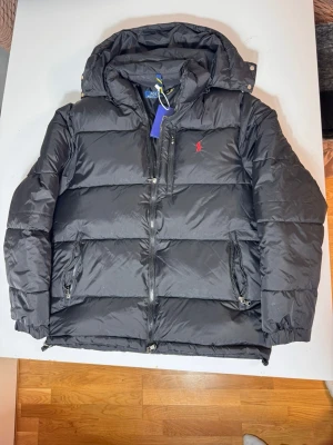 Svart pufferjacka  - Svart pufferjacka med huva, två sidofickor med dragkedja och röd broderad logga på bröstet. Jackan har en chunky dragkedja framtill och är tillverkad i polyester, perfekt för kalla dagar. Finns i storlek S och M 